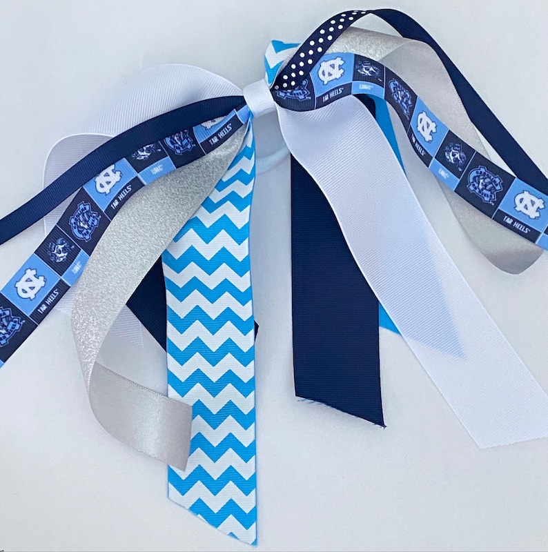 May include: A blue and white hair tie with a chevron pattern, a silver glitter ribbon, a white ribbon, a navy blue ribbon, and a ribbon with the UNC Tar Heels logo and the words "Tar Heels".