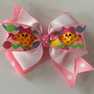May include: A pink and white hair bow with a colorful Easter chick design. The bow has white polka dots on the pink edges.