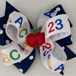 Op de afbeelding: Een wit en marineblauwe haar strik met witte stippen. De strik heeft een rode appel bedel en een wit lint met de letters A, B, C en de cijfers 1, 2, 3 erop gedrukt.