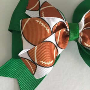 May include: A green and white hair bow with a football pattern. The bow is made of two layers of ribbon, with the top layer featuring a white background with brown footballs and the bottom layer being solid green.