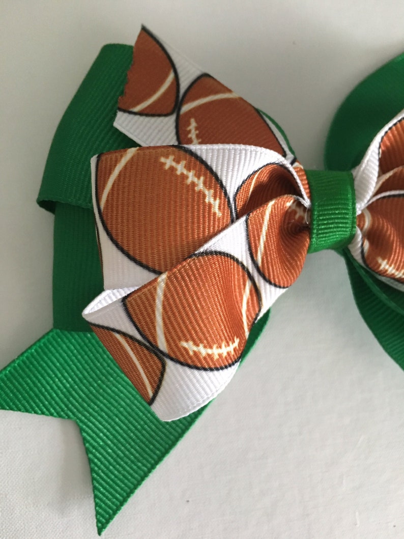 May include: A green and white hair bow with a football pattern. The bow is made of two layers of ribbon, with the top layer featuring a white background with brown footballs and the bottom layer being solid green.