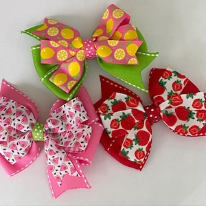 May include: Four colorful hair bows with fruit patterns. One bow features lemons on pink and green ribbons. Another has watermelon slices on pink. The other two bows have strawberries on red and white ribbons.