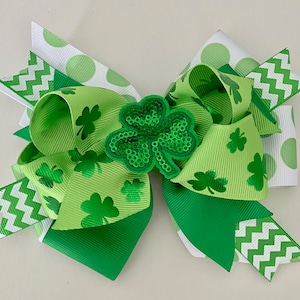 May include: A green, white, and green shamrock hair bow with a sparkly green shamrock accent. The bow is made of ribbon with a polka dot pattern, a chevron pattern, and a shamrock pattern.