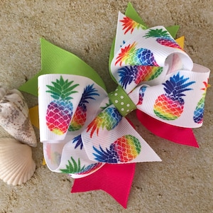 May include: A white hair bow with a rainbow pineapple print. The bow has green, pink, and white ribbon.