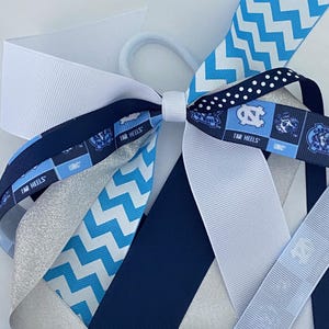 May include: A blue and white hair bow with a chevron pattern, a white and blue polka dot pattern, and a silver glitter ribbon. The bow also features a blue and white ribbon with the text "TAR HEELS" repeated on it.