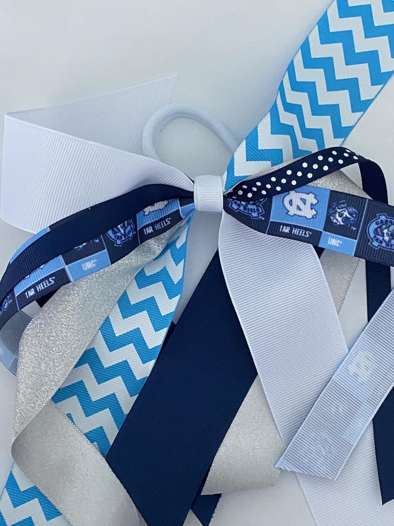 May include: A blue and white hair bow with a chevron pattern, a white and blue polka dot pattern, and a silver glitter ribbon. The bow also features a blue and white ribbon with the text "TAR HEELS" repeated on it.