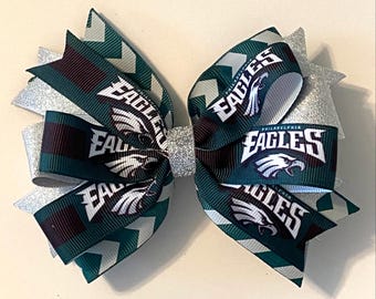 May include: Green, white, and black hair bow with a chevron pattern and the Philadelphia Eagles logo. The bow is decorated with silver glitter.