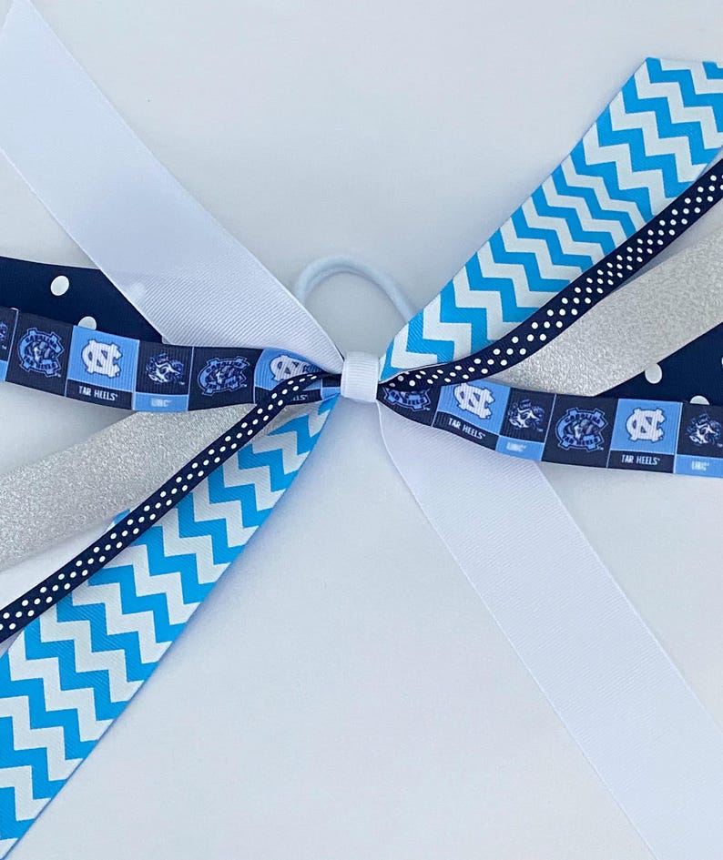 May include: A blue and white hair bow with a chevron pattern, white polka dots, silver glitter ribbon, and a blue and white ribbon with the text "Tar Heels".