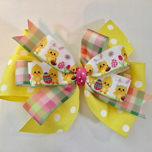 May include: A yellow, white, and pink plaid ribbon bow with a white ribbon featuring a pattern of chicks and Easter eggs. The bow is tied with a pink polka dot ribbon.
