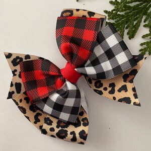 May include: A large hair bow featuring layers of red and black buffalo plaid, black and white checkered, and tan leopard print ribbons. The bow is tied with a red center knot. A sprig of greenery is in the background.