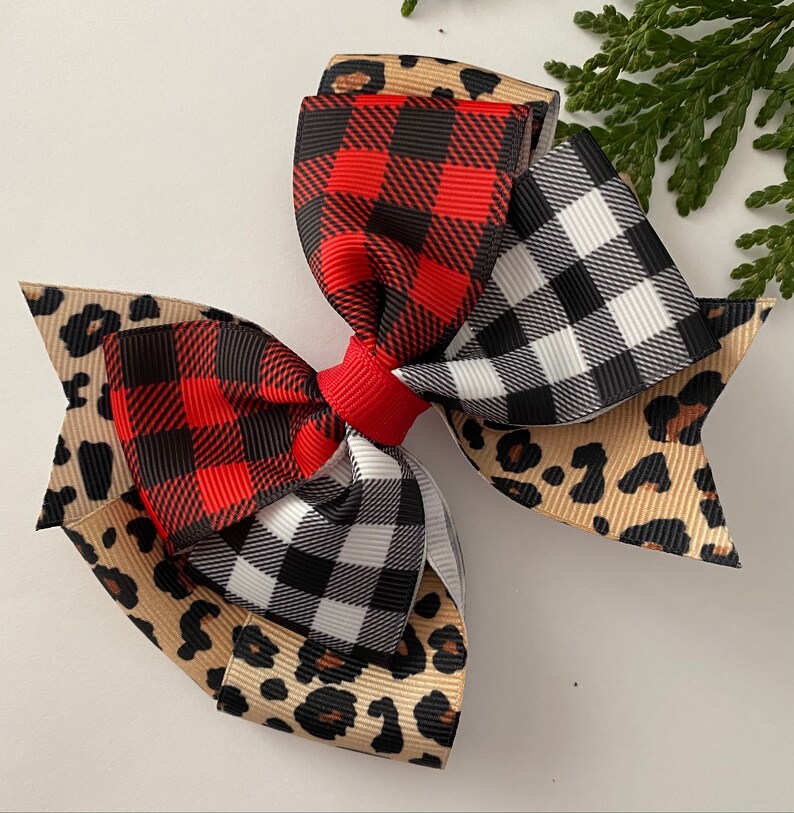 May include: A large hair bow featuring layers of red and black buffalo plaid, black and white checkered, and tan leopard print ribbons. The bow is tied with a red center knot. A sprig of greenery is in the background.