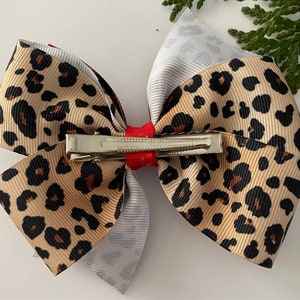 May include: A large hair bow featuring a layered design. The top layer is a tan ribbon with black leopard spots. The middle layer is a gray and white striped ribbon, and the bottom layer is a burgundy ribbon. A red ribbon and a metal clip secure the bow.