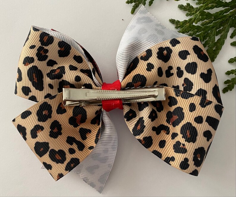 May include: A large hair bow featuring a layered design. The top layer is a tan ribbon with black leopard spots. The middle layer is a gray and white striped ribbon, and the bottom layer is a burgundy ribbon. A red ribbon and a metal clip secure the bow.