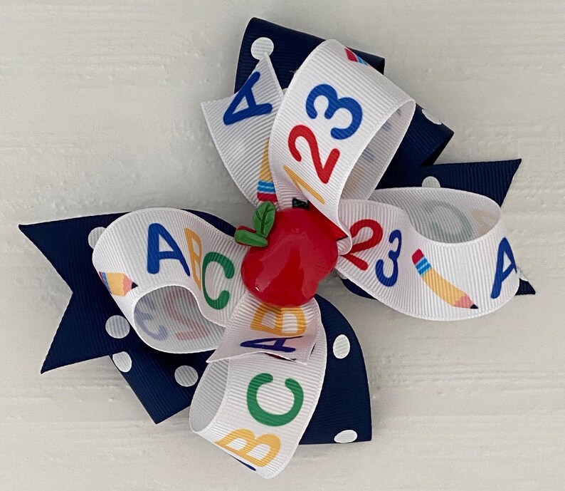 Op de afbeelding: Een wit en marineblauwe haar strik met een rode appel bedel. De strik heeft een wit lint met de letters A, B, C en de cijfers 1, 2, 3 erop gedrukt.