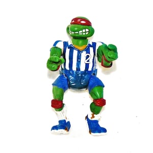 May include: A green Teenage Mutant Ninja Turtles action figure wearing a blue and white striped jersey with the number "2" on the front. The figure is wearing red knee pads and blue shoes.