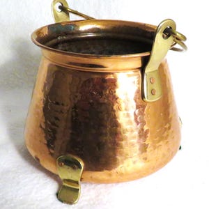 May include: A hammered copper pot with brass handles and feet. The pot has a rounded body and a wide opening.