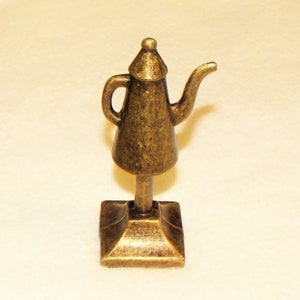 May include: A gold-colored metal teapot figurine with a square base. The teapot has a spout and a handle.