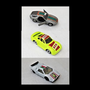 May include: Three toy cars, a silver Porsche 928, a yellow Porsche 935 with "1000" and "935" printed on it, and a white Lamborghini Countach with a red, yellow, and green Italian flag on the side.