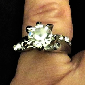 May include: A silver ring with a large, clear, round gemstone surrounded by smaller, clear gemstones.
