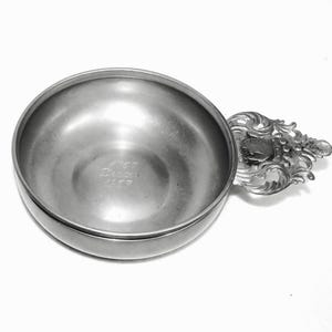 May include: A silver pewter bowl with a decorative handle. The bowl has a round shape and a smooth surface. The handle is ornate and features a crest with a grapevine design. The bowl is inscribed with the text "1767 Dresden 1817".