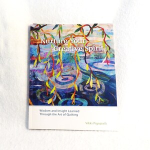 May include: A book titled "Nurture Your Creative Spirit" with colorful artwork of a tree over water. The book cover includes the text "Wisdom and Insight Learned Through the Art of Quilting" and the author's name, Vikki Pignatelli.