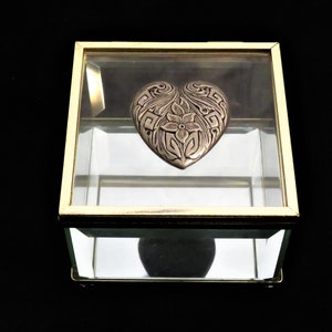 May include: A glass box with a gold trim, containing a silver heart-shaped ornament with an intricate floral design.