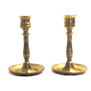 May include: A pair of gold metal candle holders with a simple design. Each holder has a round base, a tall, slender stem, and a cup-shaped top.