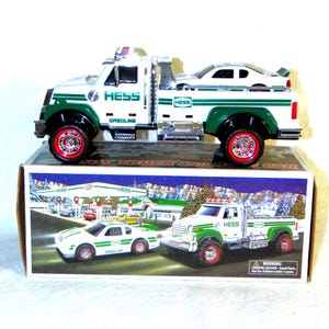 May include: A white and green Hess toy truck and car set. The truck has red wheels and the word "HESS" printed on the side. The car is white. The box features a winter scene with a gas station and other vehicles.