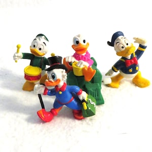 May include: Four Disney duck figurines in various poses. One plays a drum, another sits on a green throne, and a third strikes a pose. The fourth duck figure is walking with a cane and a green bag. Each figure has a unique outfit.