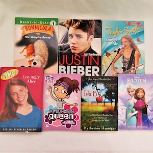 May include: A collection of eight paperback books featuring various genres, including children's fiction, biographies, and young adult fiction. The books include titles like "Bunnicula", "Justin Bieber", "Taylor Swift", "Alice", "Go Girl! Dancing Queen", "Ida B", and "Frozen".