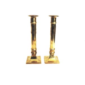 May include: A pair of gold metal candle holders with a square base and a cylindrical design.