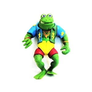 May include: A green frog action figure wearing a blue and yellow jacket, red shorts, and a gold chain. The figure has a toothy grin and large, round eyes.