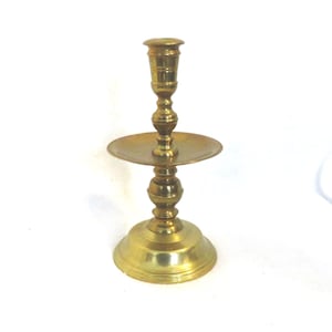 May include: A gold metal candlestick with a round base and a single candle holder. The candlestick has a decorative design with a round plate in the middle.