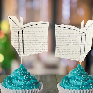 May include: Two teal blue cupcakes with white frosting and white sprinkles. Each cupcake is topped with a book page shaped like a butterfly. The text on the book pages reads "The police...back seven large, tough men growing impatient." and "...here the Cache is, so we try to prevent him from getting to it."