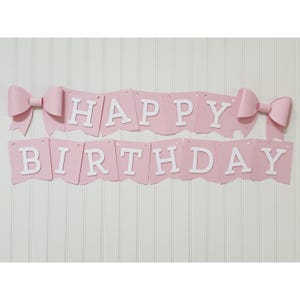 May include: A pink banner with the words "HAPPY BIRTHDAY" in white letters. The banner is decorated with two large pink bows, one on each end. The banner is hanging against a white wall.