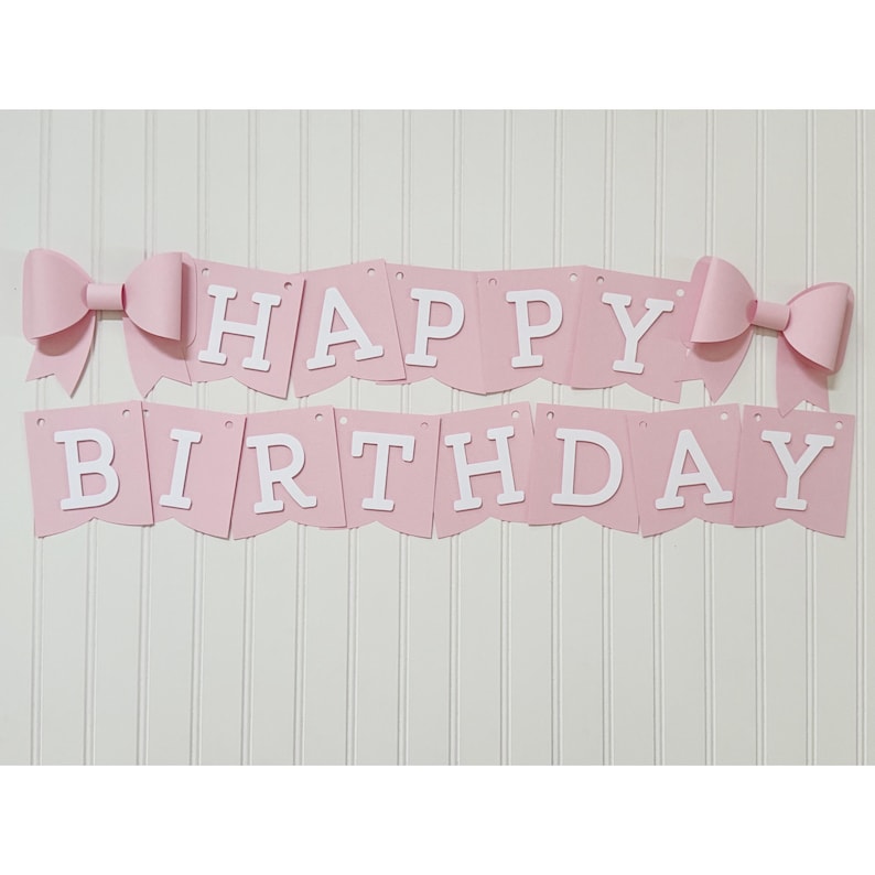 May include: A pink banner with the words "HAPPY BIRTHDAY" in white letters. The banner is decorated with two large pink bows, one on each end. The banner is hanging against a white wall.