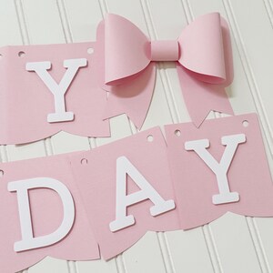 May include: A pink paper banner with the word "DAY" and "Y" spelled out in white letters. A large pink paper bow is attached to the banner. The banner is designed for a birthday celebration.