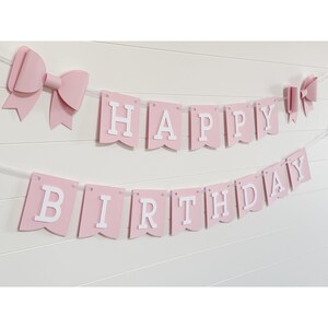 May include: Pink "Happy Birthday" banner with white lettering and two pink bow decorations. The banner is strung on a white string and is hanging on a white wall.