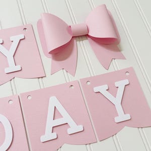 May include: A pink paper banner with white letters spelling "DAY" and a large pink bow. The banner pieces are rectangular with a scalloped bottom edge and small holes at the top for hanging. The bow is three-dimensional.