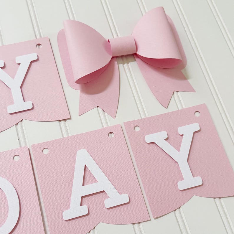 May include: A pink paper banner with white letters spelling "DAY" and a large pink bow. The banner pieces are rectangular with a scalloped bottom edge and small holes at the top for hanging. The bow is three-dimensional.