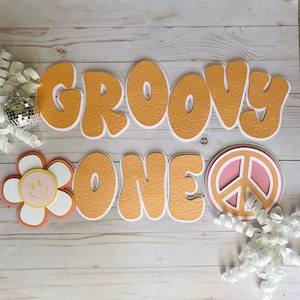 May include: Groovy One banner with orange letters and peace sign pattern. The letters are in a retro font and the banner is decorated with a pink and white peace sign and a yellow flower with a smiley face.