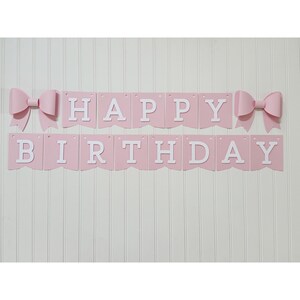 May include: A pink banner with the words "HAPPY BIRTHDAY" in white letters. The banner is decorated with two pink bows, one on each side. The banner is hanging on a white wall.