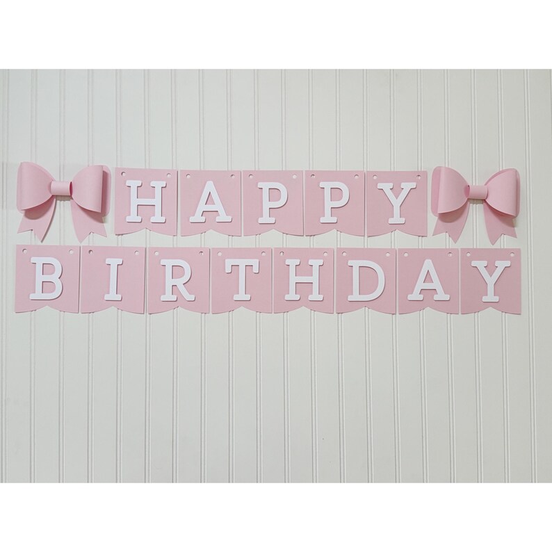 May include: A pink banner with the words "HAPPY BIRTHDAY" in white letters. The banner is decorated with two pink bows, one on each side. The banner is hanging on a white wall.