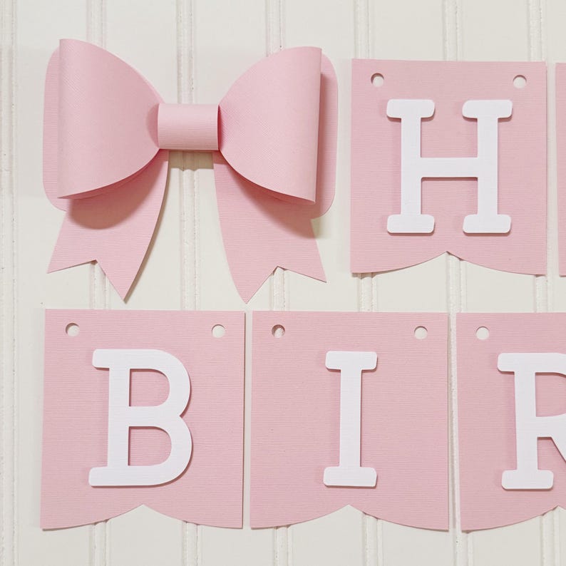 May include: A pink paper bow and a banner spelling out "BIRTHDAY" in white letters. The banner is made of pink paper pennants with white letters. The bow is large and made of layered pink paper.