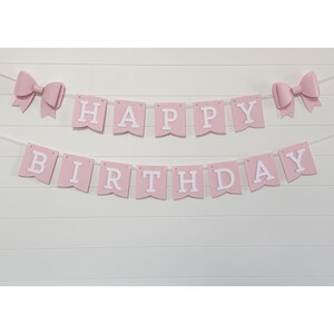 May include: Pink "Happy Birthday" banner with white lettering and two pink bow accents. The banner is strung on a white string and is displayed against a white, horizontal-slat background. Perfect for a birthday party.