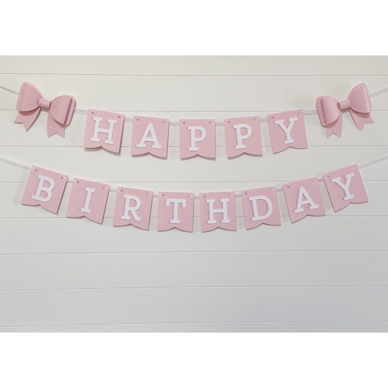 May include: Pink "Happy Birthday" banner with white lettering and two pink bow accents. The banner is strung on a white string and is displayed against a white, horizontal-slat background. Perfect for a birthday party.