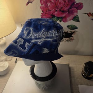 May include: Blue bucket hat with the word "Dodgers" in white script. The hat has a blue and white pattern and a dark blue brim. The hat is displayed on a white mannequin head.