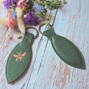 May include: Two leaf-shaped keychains made of green material, each attached to a bronze-colored metal ring. One keychain features a colorful, patterned bee illustration, while the other displays decorative stitching in a leaf vein pattern.