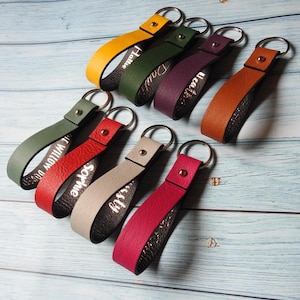 Op de afbeelding: Een verzameling gepersonaliseerde leren sleutelhangers in diverse kleuren, waaronder geel, groen, paars, oranje, rood, beige en roze. Elke sleutelhanger heeft een zilveren sleutelring en een witte naam. Gepresenteerd op een lichtblauwe houten ondergrond.