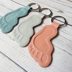 Puede incluir: Tres llaveros de cuero con forma de huellas de pies descalzos. Los llaveros son de color azul claro, gris y melocotón.